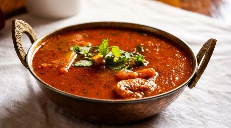 Indian curry in a copper bowl with handles, garnished with fresh cilantro on a white tablecloth.