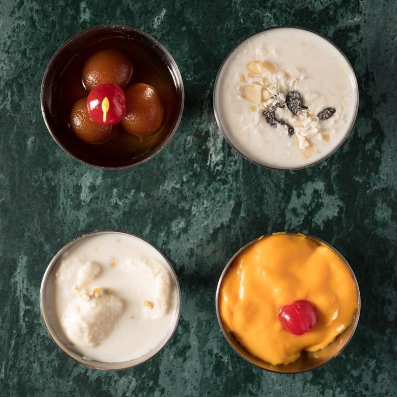 Four bowls of Indian desserts: gulab jamun, kheer, rasmalai, and mango shrikhand on a green marble surface.