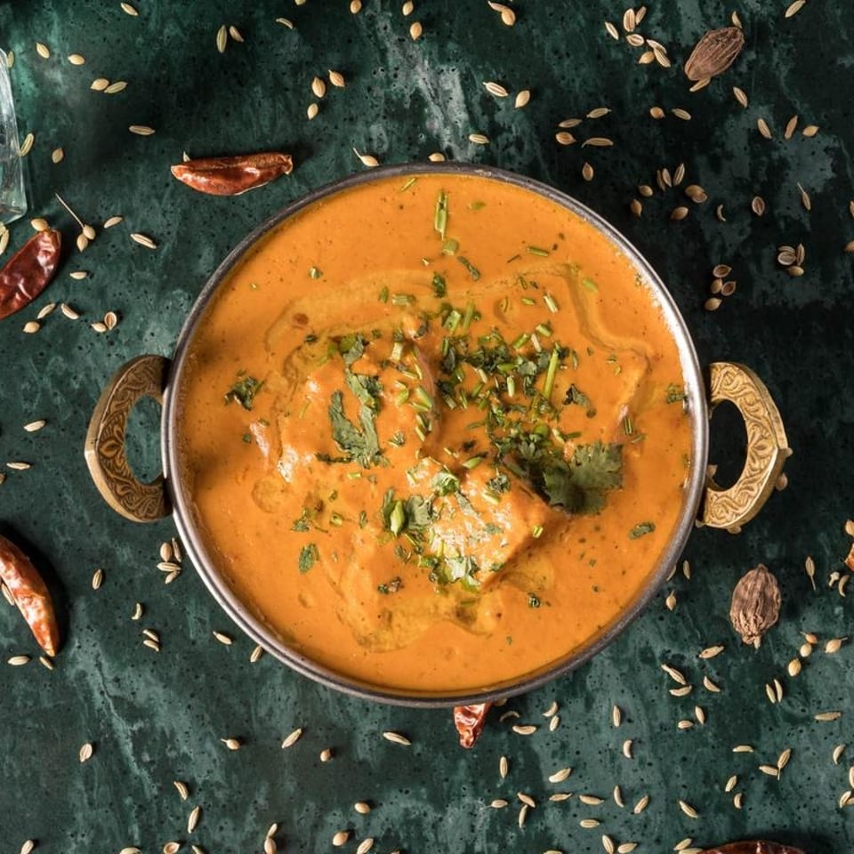 A bowl of Indian butter chicken curry with cilantro on a dark green surface with spices.