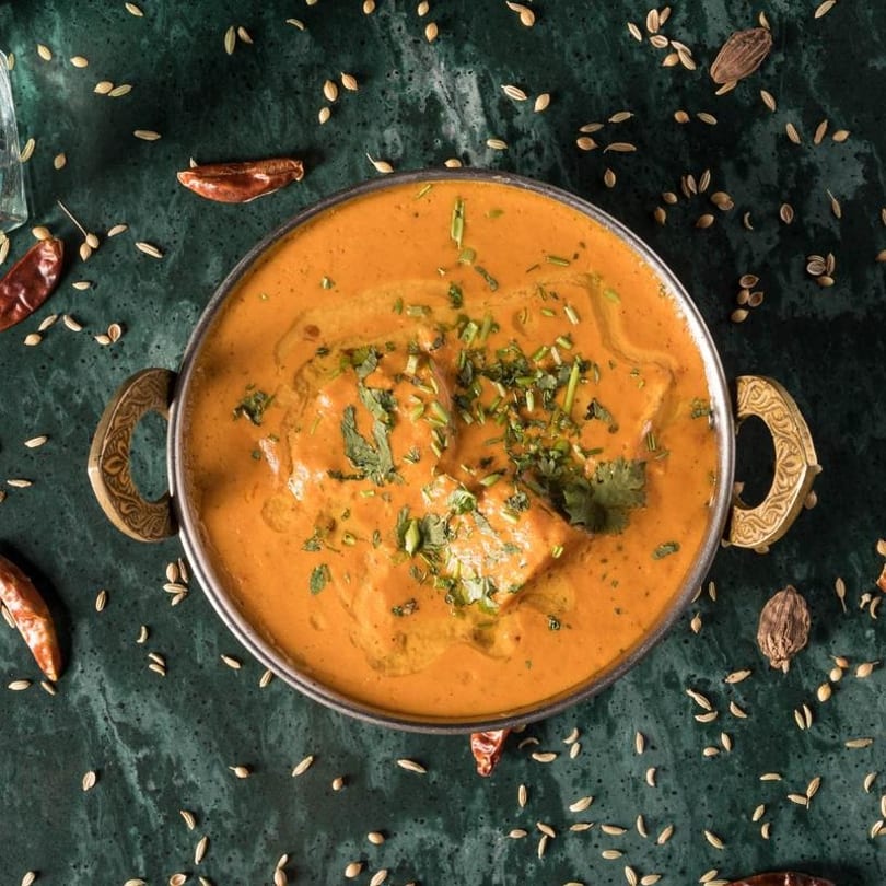 A bowl of Indian butter chicken curry with cilantro on a dark green surface with spices.