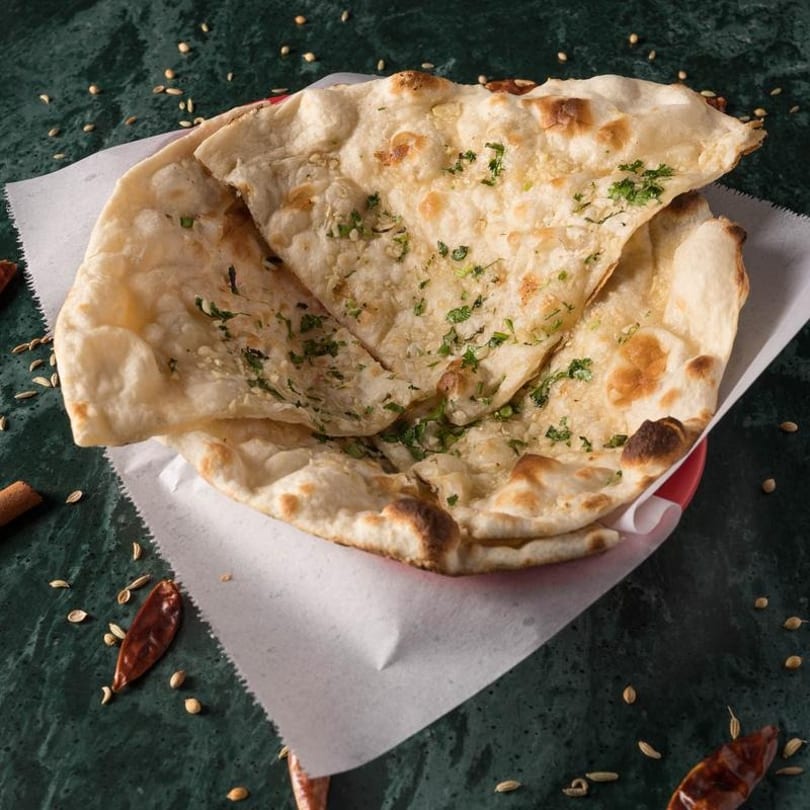 Garlic naan bread, garnished with cilantro, served in a red basket on a dark green surface.