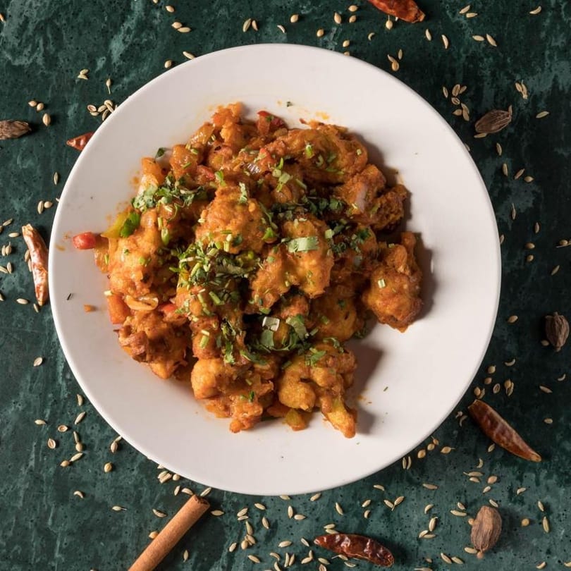 Spicy cauliflower dish garnished with fresh cilantro on a white plate, surrounded by spices.