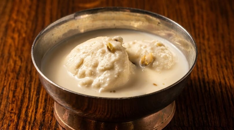 Two ras malai pieces with pistachios in a silver bowl filled with creamy milk on a wooden table.