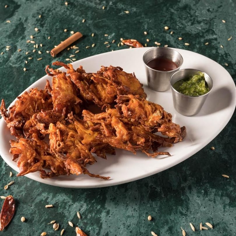 Crispy fried onion bhaji on a white plate with green and red chutneys, spices scattered around.