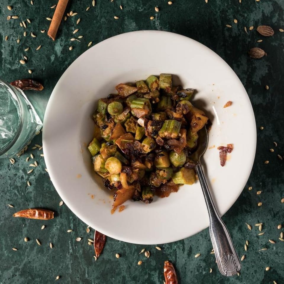 A white bowl of cooked okra and onions with a spoon, on a green table with spices.