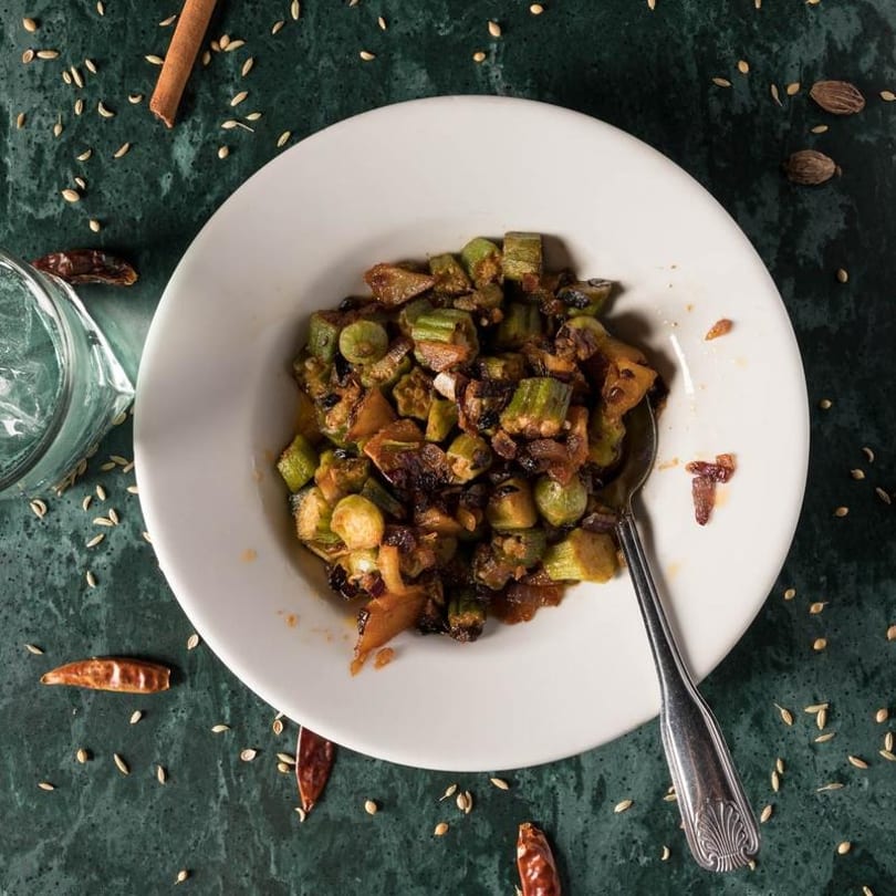 A white bowl of cooked okra and onions with a spoon, on a green table with spices.