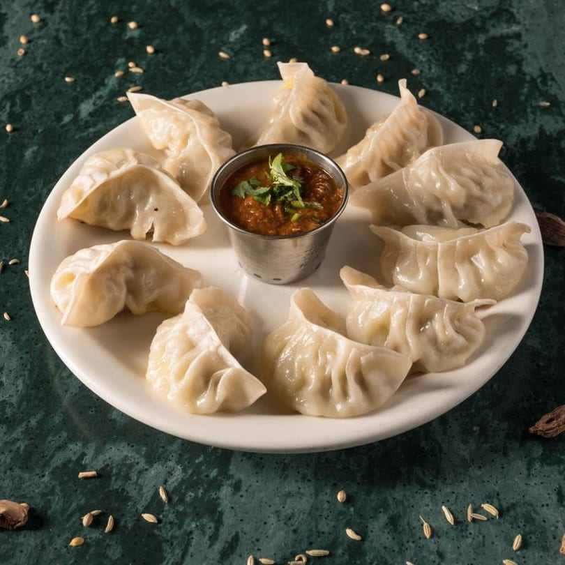 A white plate of steamed dumplings arranged around a small metal cup of red dipping sauce.
